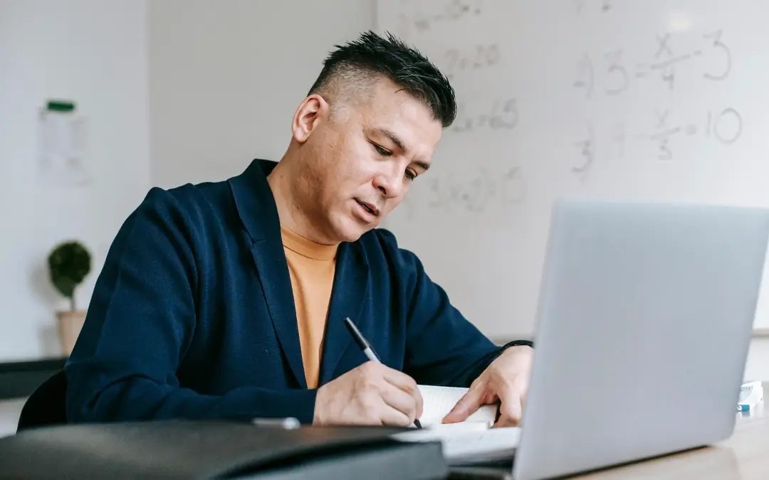 homem escrevendo em papel na mesa com laptop à frente