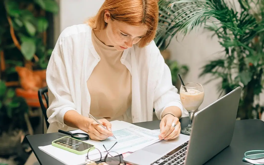 mulher ruiva analisando papéis na mesa com laptop à frente
